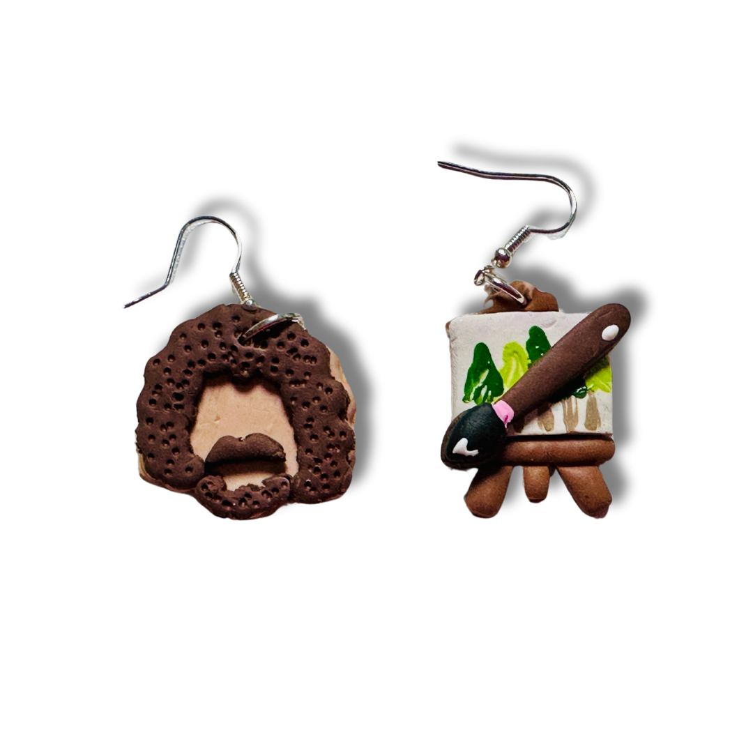 Handmade Bob Ross Style Polymer Clay Earrings. The earring on the left is a 1x 1 inch Bob Ross Head shaped with beige skin, brown hair, beard, and mustache. The earring on the right is a 0.75 x 1 inch paint canvas with a paintbrush on top. The canvas shows a brown easel holding a white canvas with hand painted green trees. On top of the canvas is a brown paint brush with black brush and shine of white to reflect the lighting. These earrings have nickel free silver hooks.
