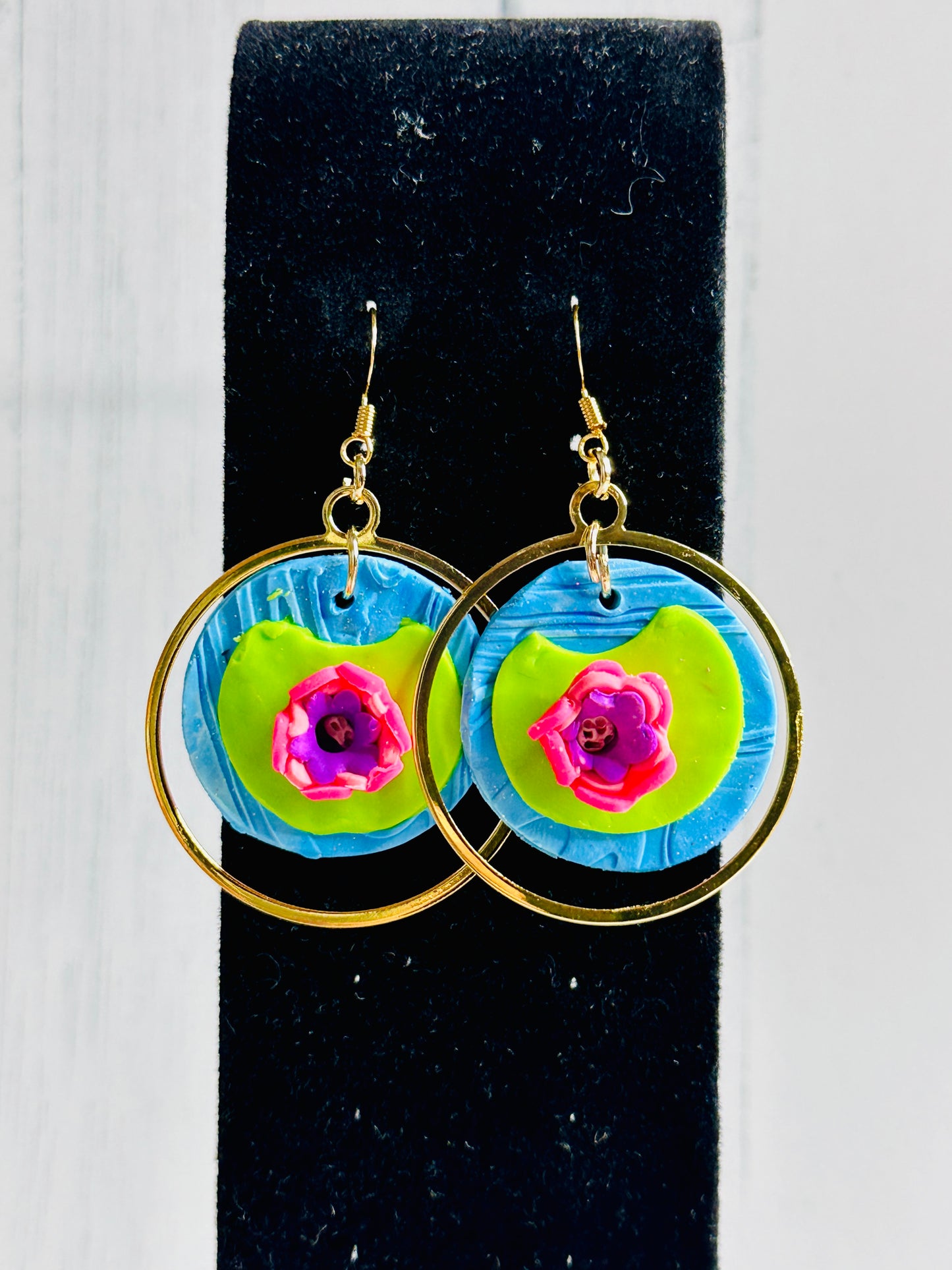 A closeup of the Monet Water Lily Earrings on a black stand with a bright white wall behind it.
