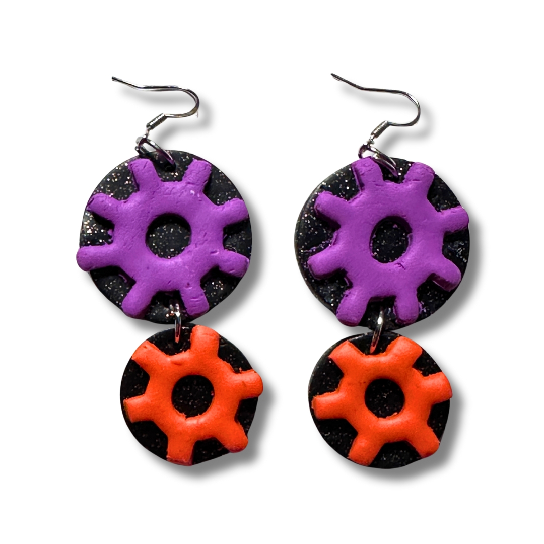 These pair of STEM earrings resting on a white background show the 1.25in purple round gear on a glittery black circle background. Hooked along with a smaller orange round gear at 1 inch on a glittery black circle background. The earrings also have nickel free silver hoops.