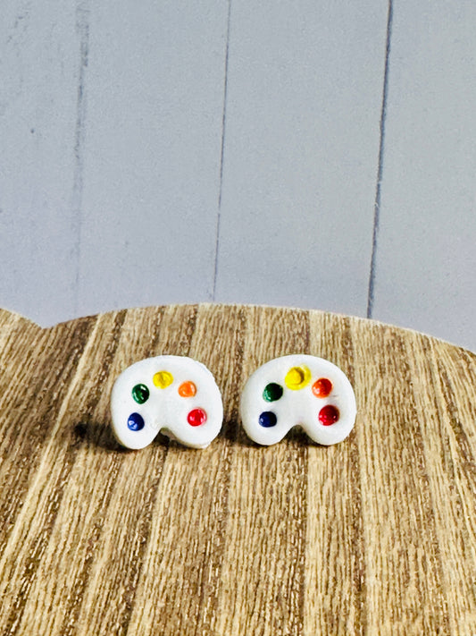 A pair of art palette stud earrings on a wooden table.  The palette is white and has dots of blue, green, yellow, orange, and red paint.