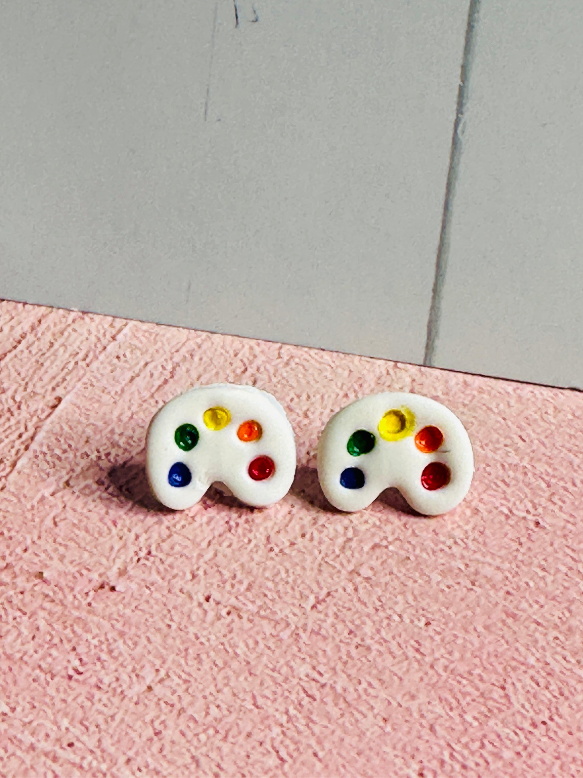 A pair of art palette stud earrings on a pink table in front of a white wall.  The palette is white and has dots of blue, green, yellow, orange, and red paint.