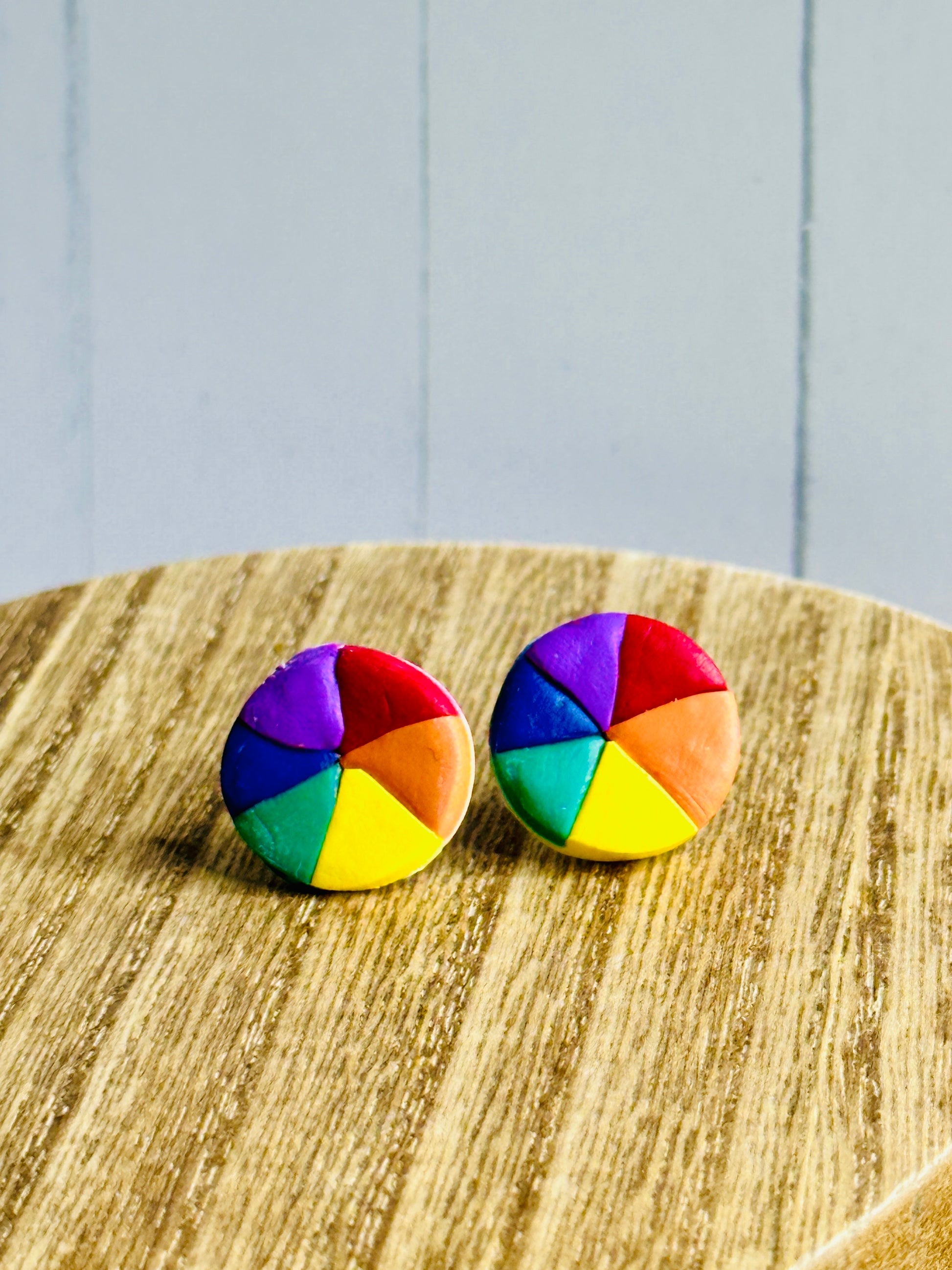 A pair of polymer clay earrings shaped like color wheels for artists and art teachers sitting on a wooden table.
