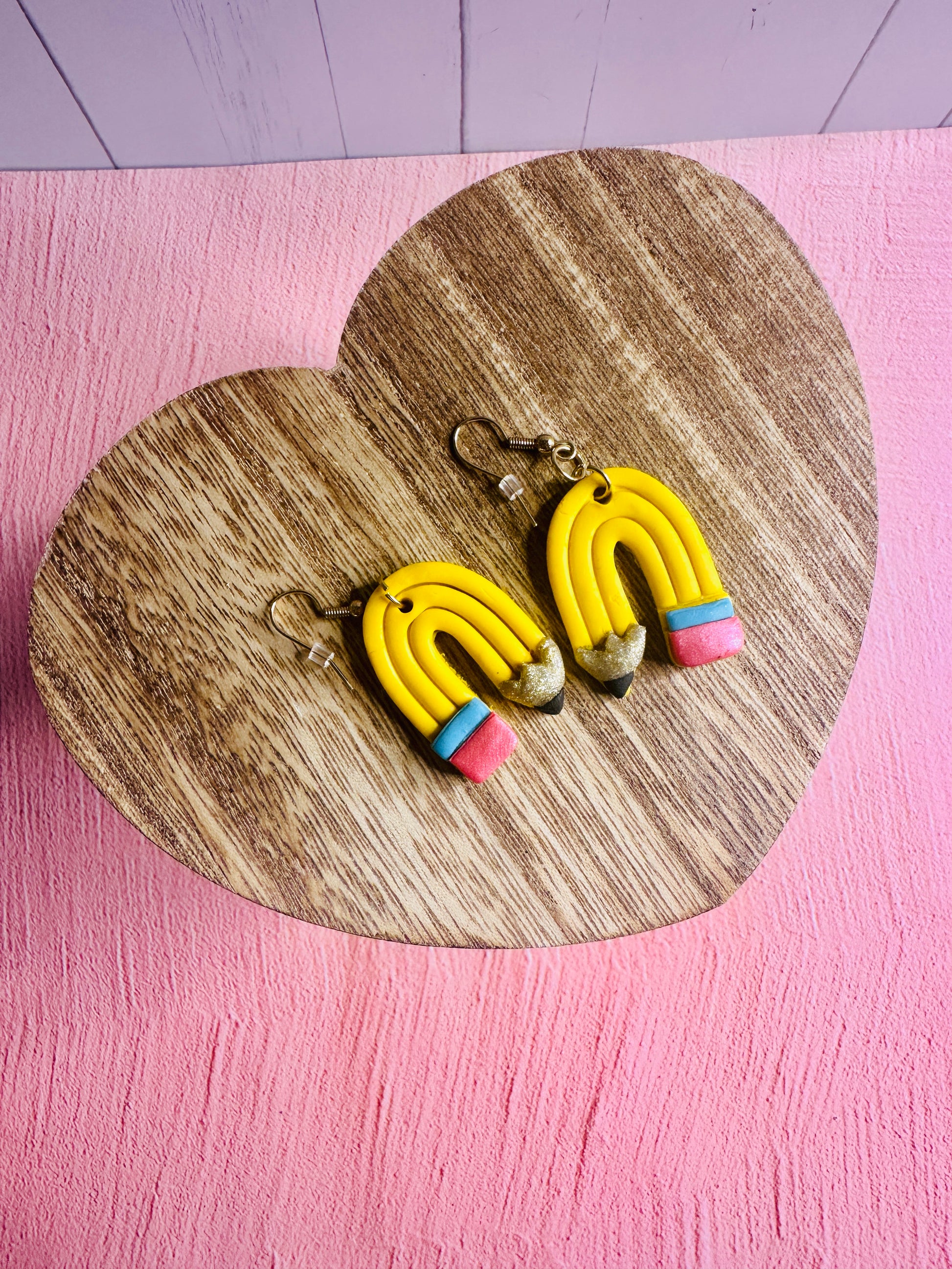 The Curved Pencil Earrings on a wooden table with a pink background.