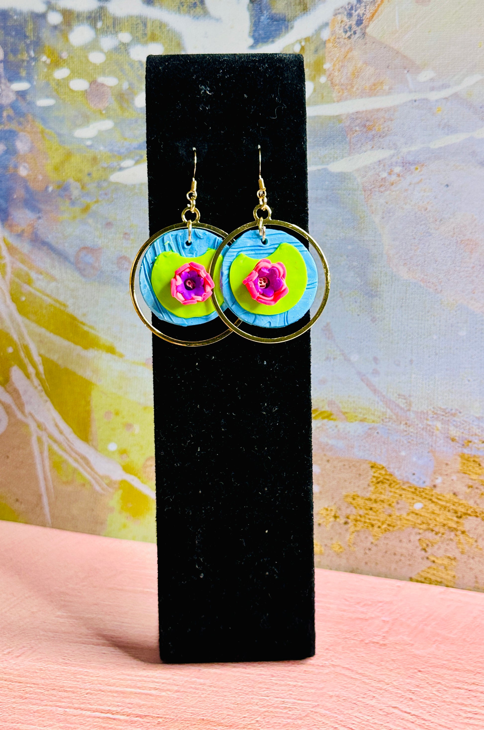 A pair of Monet Water Lily Earrings on a tall, black earring stand.  A colorful, abstract all is behind the stand.