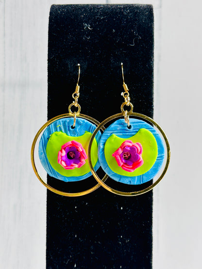 A closeup of the Monet Water Lily Earrings on a black stand with a bright white wall behind it.