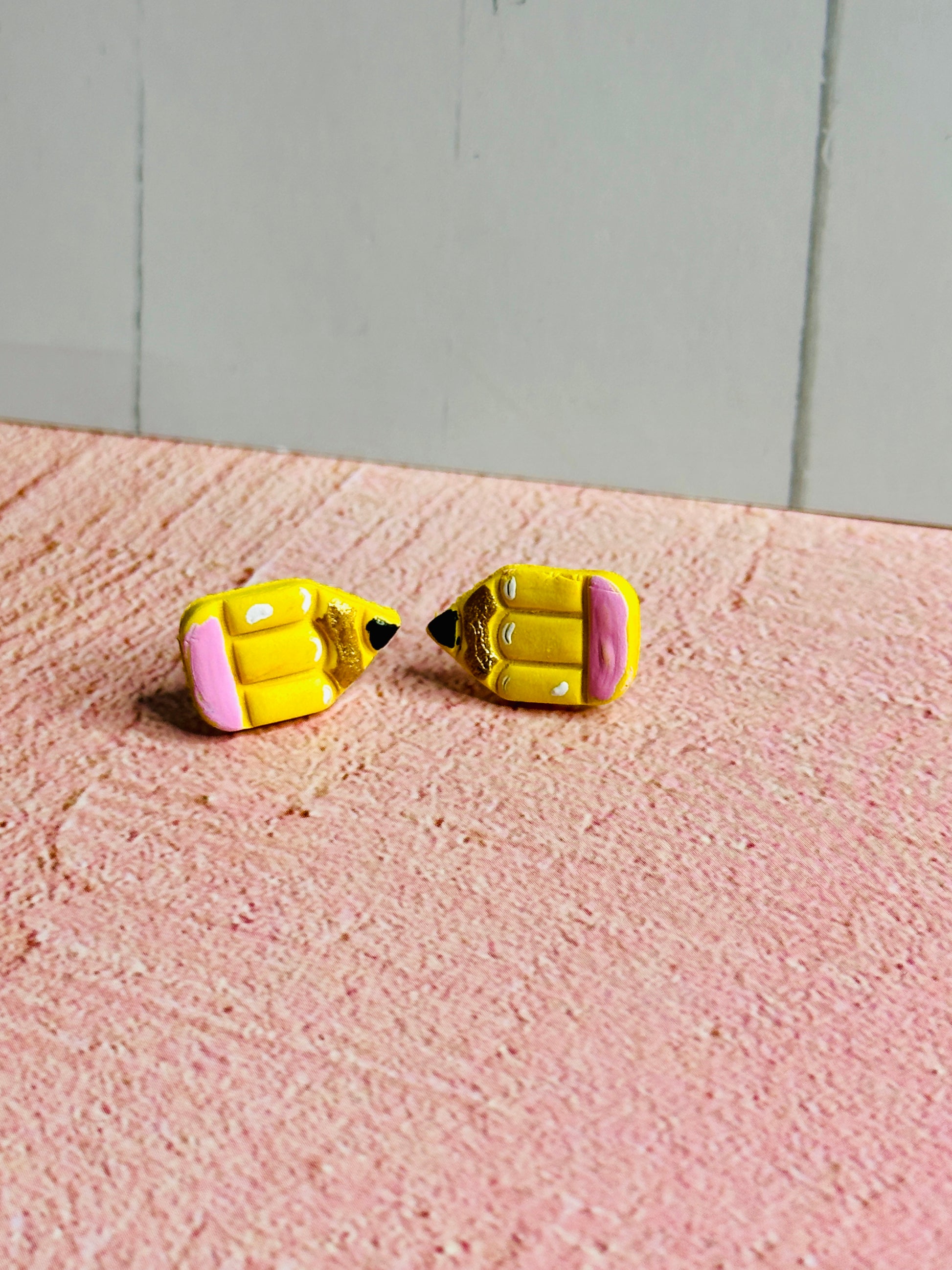 Am angled view of a set of pencil stud earrings on a pink table  The stud is yellow with a pink eraser, white shadows, gold colored wood, and black pencil tip.