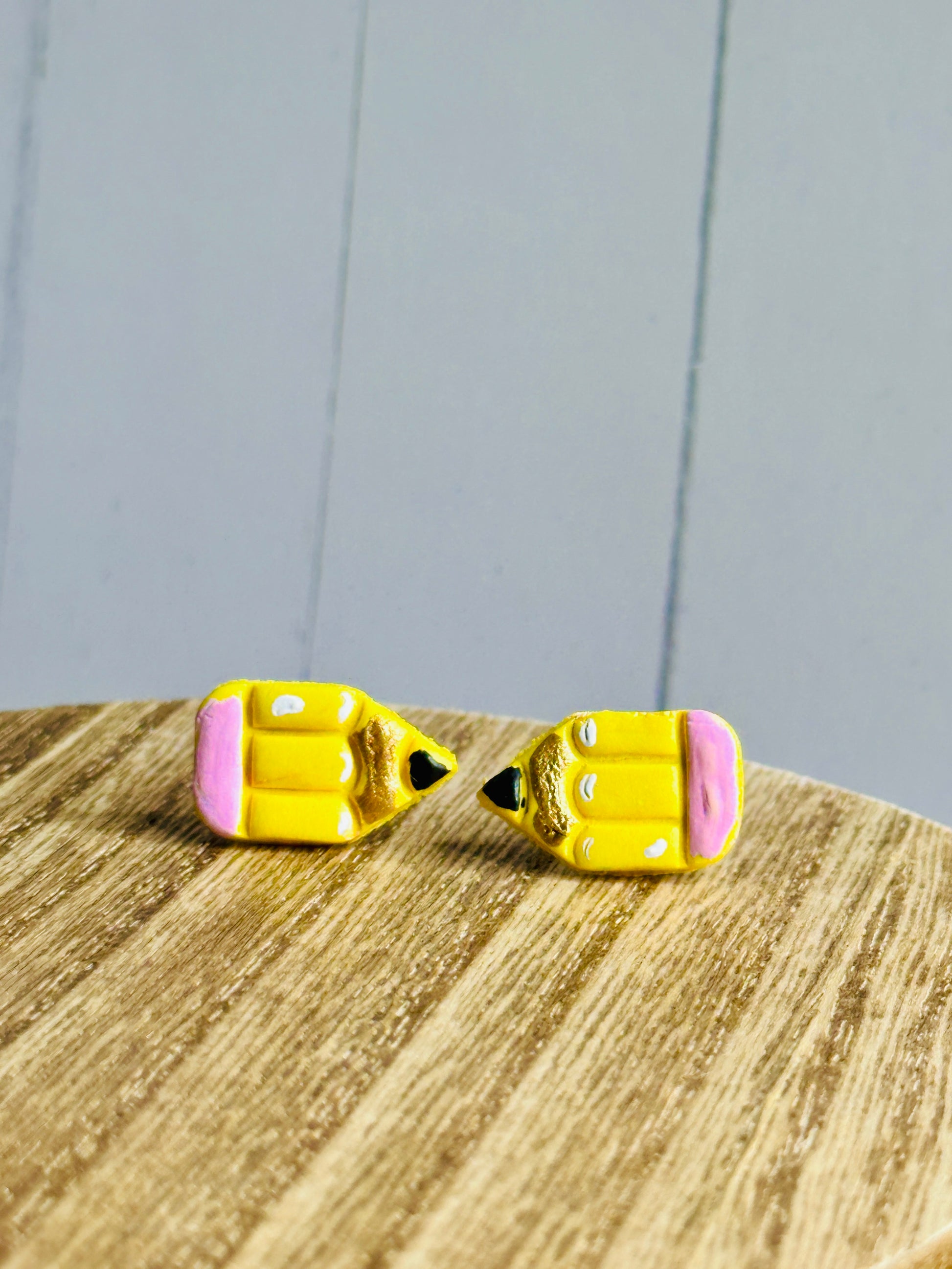 A set of pencil stud earrings on a wooden table  The stud is yellow with a pink eraser, white shadows, gold colored wood, and black pencil tip.